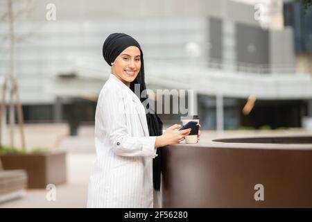 Lächelnde arabische Frau, die mit einem wiederverwendbaren Becher an der Rückhaltewand steht Und Smartphone Stockfoto