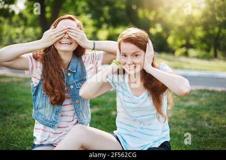 Familienfoto von zwei schönen Rotschopf Mädchen mit Sommersprossen herumtollend, während auf Gras im Park sitzen während Picknick mit besten Freunden, Augen deckend Stockfoto