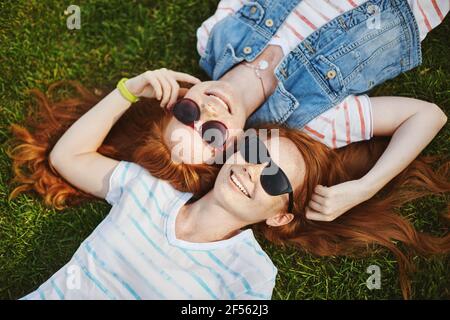 Blick des Glücks. Zwei sonnige und glückliche Zwillingsschwestern mit Ingwerhaaren, die auf grünem Gras liegen und Köpfe berühren, mit Haarsträhnen spielen und lächeln Stockfoto