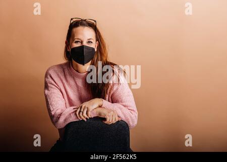 Junge Frau, die eine schützende Gesichtsmaske trägt, starrt an, während sie ansteht Beigefarbener Hintergrund Stockfoto