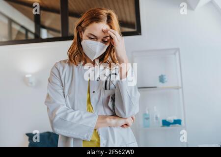 Arzt trägt eine schützende Gesichtsmaske und steht mit dem Kopf in den Händen In der Klinik Stockfoto