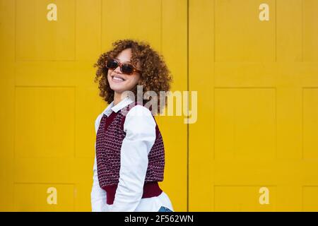 Lächelndes Mädchen im Teenageralter mit Sonnenbrille, das gegen die gelbe Tür steht Stockfoto