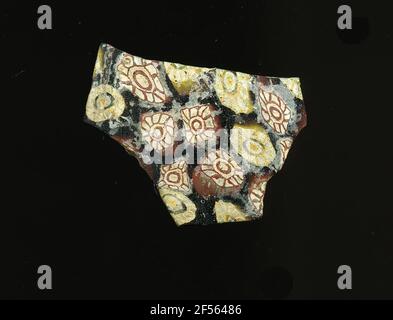 Fragment eines Gefäßes. Eine niedrige Krümmung des Glassfragmentes ermöglicht die Zuordnung zu einem Gefäß, möglicherweise einer Schale. Der schwarze Scherbe zeigt zwei verschiedene Arten von Blumen in der Millefiori-Technik: Die eine besteht aus einem weißen Punkt mit roten, weißen und roten Rand, um die roten Punkte kreisförmig als Blütenblätter zu gruppieren, die anderen bestehen aus einem gelben Punkt mit weißen, schwarzen und gelben Rand, um die weißen Punkte mit gelben Kontur die Blütenblätter darstellen. Für die Herstellung wurden verschiedenfarbige Gläser zu Rohren und Stäben geformt. Diese wurden komprimiert und erhitzt, was zu einem langen Balken führte. Die Glasscheiben hatten ​​now anders Stockfoto