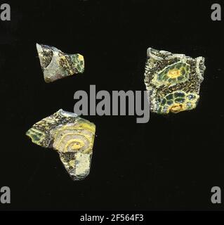 Drei Fragmente eines Gefäßes. Eine niedrige Krümmung der drei kombinierten Glassplitter ermöglicht die Zuordnung zu einem Gefäß. Sie zeigen auf dunkelviolettem Hintergrund zwei Blütenformen in Millefiori-Technik: Eine besteht aus einer gelben Spirale, die in einem gelben Zentrum entspringt. Die anderen bestehen aus einem grünen Punkt mit gelber Kontur, um die gelb umrandeten grünen Blütenblätter zu liegen. Für die Herstellung wurden verschiedenfarbige Gläser zu Rohren und Stäben geformt. Diese wurden komprimiert und erhitzt, was zu einem langen Balken führte. Die Glasscheiben hatten verschiedene amorphe oder florale Muster. ​​now Danach waren mehrere dieser Thrombozyten mer Stockfoto
