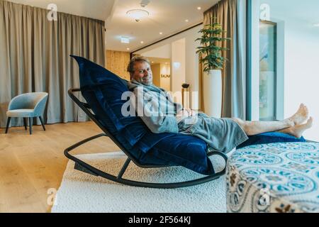 Mann im Bademantel, der sich auf einem Schaukelstuhl im Hotelzimmer entspannt Stockfoto