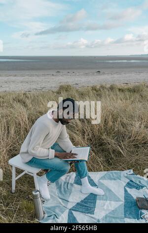 Junger Mann liest Buch, während er am Strand auf dem Hocker sitzt Stockfoto