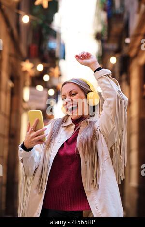 Fröhliche Frau mit Mobiltelefon, die tanzt, während sie Musik hört Kopfhörer in der Gasse Stockfoto