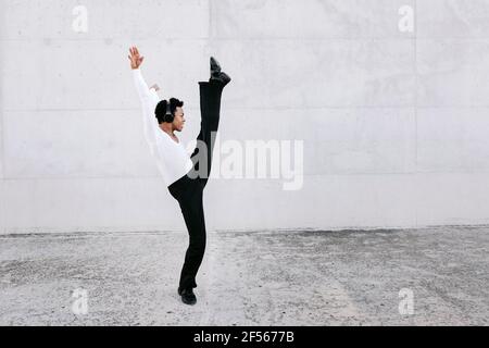 Flexibler männlicher Tänzer, der sein Bein streckt, während er den urbanen Tanz an der weißen Wand übt Stockfoto