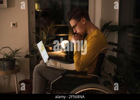 Konzentrierter Mann im Rollstuhl mit Laptop, der spät arbeitet Sitzen auf Rollstuhl im Wohnzimmer Stockfoto