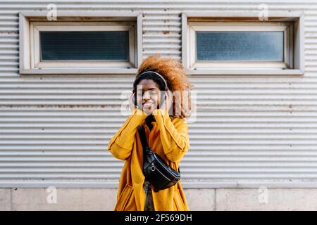 Lächelnde Afro-Frau mit geschlossenen Augen und Musik über Funk hören Kopfhörer, während sie gegen die Wand stehen Stockfoto