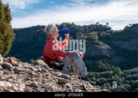 Männlicher Wanderer, der Wasser trinkt, während er auf dem Berg in Sant Llorenc del Munt i l'Obac, Katalonien, Spanien sitzt Stockfoto