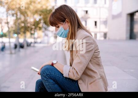 Junge Frau mit schützender Gesichtsmaske mit Smartphone, während Draußen sitzen Stockfoto