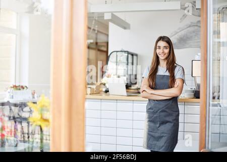 Junge Unternehmerin, die ein erfolgreiches kleines Café betreibt. Stehen vor der Kaffeemaschine mit gekreuzten Armen. Stockfoto