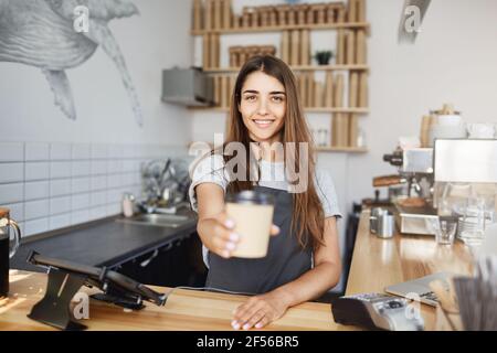 Junge Barista, die dem Gast eine Tasse Cappucino oder spät einen Blick auf die Kamera gab. Frischen Kaffee für guten Geschmack zubereiten. Stockfoto