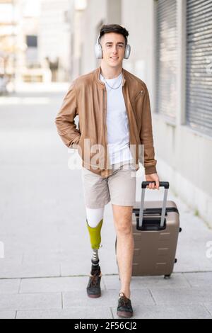 Hübscher junger Mann mit Behinderung, der Gepäck auf dem Fußweg zieht Stockfoto