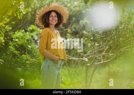Lächelnde Frau im Strohhut schaut weg, während sie im Stehen steht Garten Stockfoto