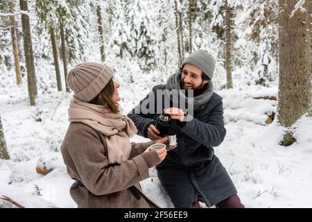 Junges Paar lächelt, während es im Winter im Wald Kaffee getrunken hat Stockfoto