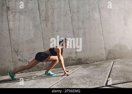 Junge Läuferin bereitet sich darauf vor, bei ihrem täglichen Urban Workout nach vorne zu joggen. Betonwände umgeben tan Mädchen. Stockfoto