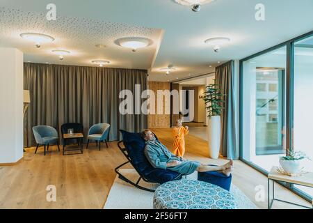 Mann, der sich auf einem Schaukelstuhl entspannt, während der Junge im Hotel gestikelt hat Zimmer Stockfoto