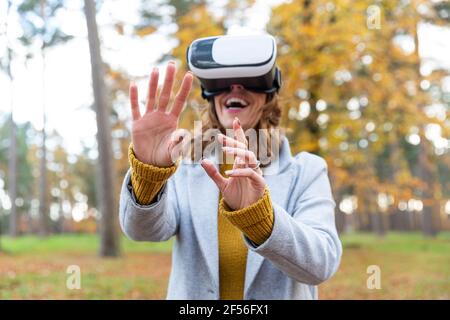 Lächelnde Frau mit Virtual-Reality-Headset, die sich im Stehen die Hände streckt Im Wald Stockfoto