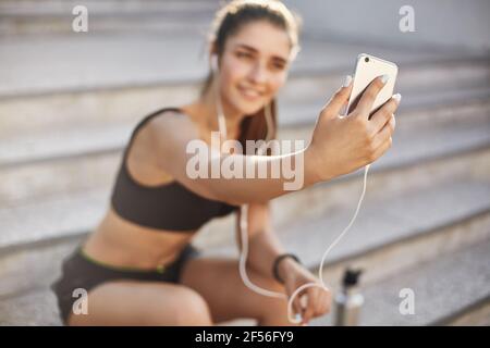Verschwommene Aufnahme mit Fokus auf Smartphone, attraktive fröhliche Sportlerin sitzen im Freien Treppe, hören Musik in kabelgebundenen Kopfhörer, Selfie Stockfoto