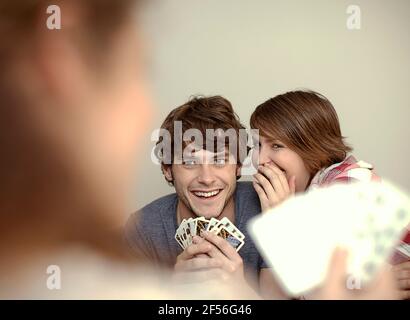 Frau flüstert dem jungen Mann ins Ohr, während sie mit Karten spielt Freund zu Hause Stockfoto