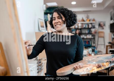 Lächelnde Künstlerin, die auf Leinwand malt, während sie die Palette anhält Kunststudio Stockfoto