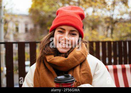 Junge Frau mit Strickmütze und Schal lächelnd im Sitzen Im Straßencafé Stockfoto