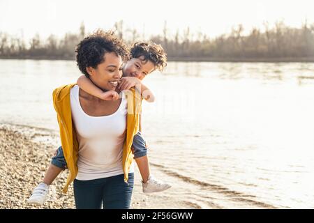 Lächelnde junge Frau Huckepack Junge beim Spaziergang am Seeufer Stockfoto