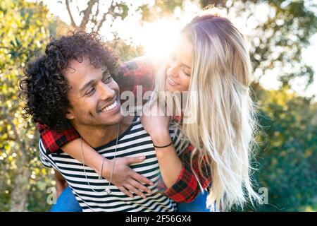 Lächelnder Mann Huckepack verspielte Frau, während Sie vor oder stehen Hinterhof Stockfoto