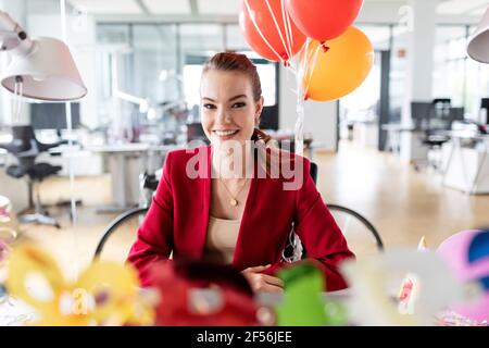 Lächelnde junge Geschäftsfrau, die während der Geburtstagsfeier im Büro sitzt Stockfoto