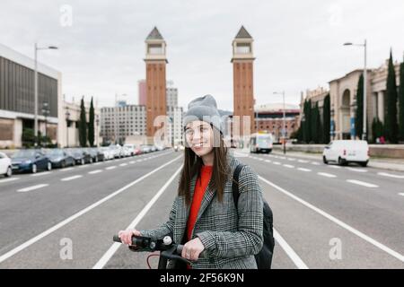 Lächelnde Frau mit Elektro-Roller auf der Straße gegen stehen Torres Venecianes in der Stadt Stockfoto