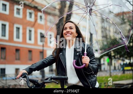 Frau, die einen Regenschirm hält, während sie auf der Straße in der Stadt Fahrrad fährt Stockfoto