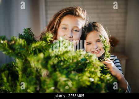 Lächelnde Mädchen, die sich zu Hause hinter dem Weihnachtsbaum verstecken Stockfoto