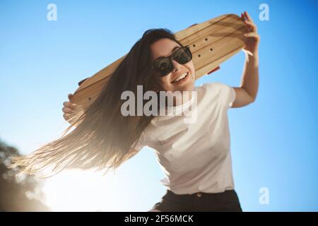 Low-Angle Schuss fröhlich glücklich junge Brünette Frau genießen Reiten Skateboard, Skater Mädchen verbringen Wochenenden Skatepark, halten hölzerne Penny Board hinter dem Kopf Stockfoto