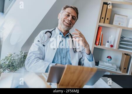 Männlicher Mitarbeiter im Gesundheitswesen, der über einen Videoanruf mit dem Patienten spricht Digitales Tablet beim Sitzen in der Arztpraxis Stockfoto