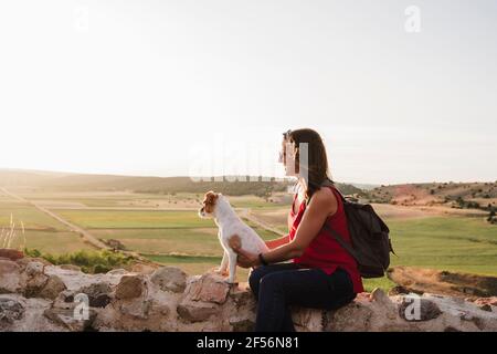 Lächelnde Frau, die Hund hält, während sie auf Felsen am Himmel sitzt Stockfoto