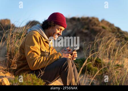 Lächelnder Mann mit Strickmütze und Smartphone im Sitzen Durch Gras Stockfoto