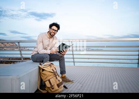 Lächelnder Geschäftsmann hält Tagebuch, während er auf der Bank am Pier sitzt Stockfoto