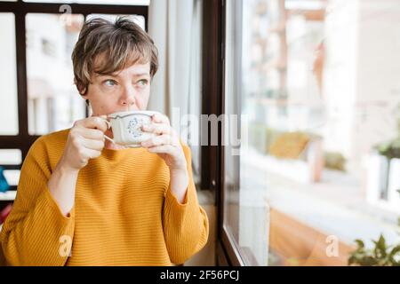 Reife Frau trinkt Kaffee, während sie zu Hause durch das Fenster schaut Stockfoto