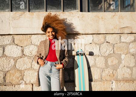 Lächelnde Frau mit Skateboard, die an sonnigen Tagen gegen das Gebäude steht Stockfoto