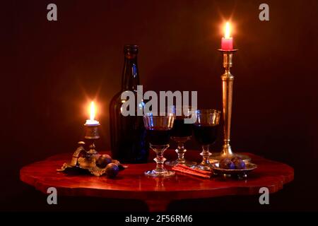 Studioaufnahme einer Flasche Rotwein, drei gefüllte Weingläser, Pflaumen, salzige Brezeln und Kerzen, die in antiken Kerzenhaltern auf einem kleinen Couchtisch brennen Stockfoto