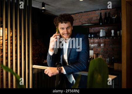Lächelnder männlicher Unternehmer, der Anzug trägt, während er auf dem Smartphone spricht Lehnt sich am Geländer im Café an Stockfoto