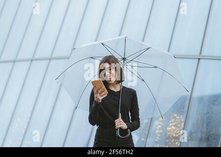 Fröhliche junge Frau, die einen Regenschirm hält, während sie ein Smartphone benutzt Vorderseite des Glasgebäudes Stockfoto