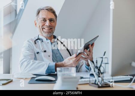 Männlicher Arzt, der eine digitale Tablette verwendet, während er in der Arztpraxis sitzt Stockfoto