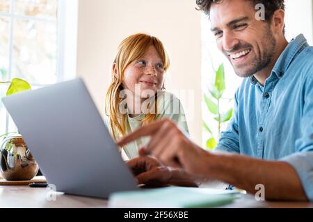 Lächelndes rothaariges Mädchen, das den Vater ansieht, der auf einem Laptop arbeitet Wohnzimmer Stockfoto