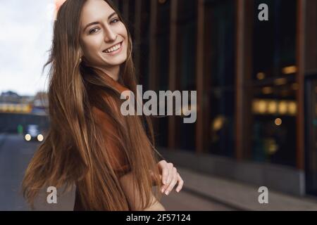 Frauen und Geschäftskonzept. Glückliche romantische junge kaukasische Frau in braunem Kunstlederkleid, drehen Sie sich nach hinten, um die Kamera zu lächeln, Wind weht auf die Haare Stockfoto