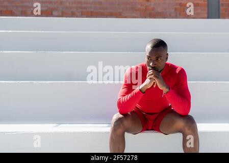 Vorderansicht junger afroamerikanischer schwarzer Sportler, der auf weißen Treppen in einem Stadion sitzt, mit verbundenen Händen, die mit ernstem fokussierten Gesicht wegschauen. Stockfoto