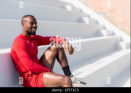 Schwarzer afrikanischer Athlet in Sportswear, der auf einer Treppe sitzt, mit lächelndem, glücklichen Gesicht, das wegschaut. Stockfoto