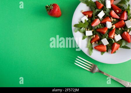 Vegetarischer Salat mit Erdbeere, Rucola, Weichkäse und Pinienkernen auf der weißen Platte auf dem grünen Hintergrund.Draufsicht. Stockfoto